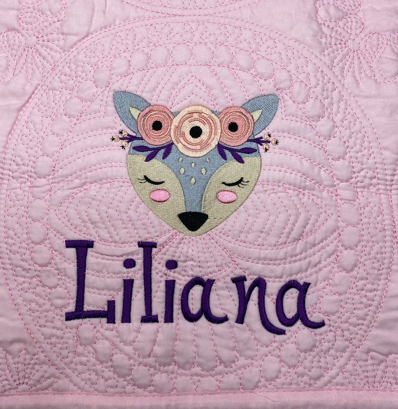 K&ouml;nnte beinhalten: Eine rosa gesteppte Babydecke mit einem gestickten Hirsch mit Blumen und dem Namen "Liliana" in lila Stickerei.