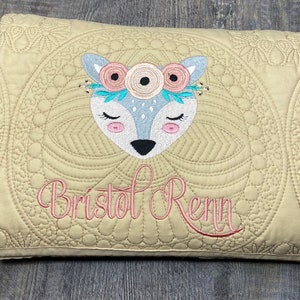 K&ouml;nnte beinhalten: Eine beige gesteppte Babydecke mit einem wei&szlig;en Hirsch, der darauf gestickt ist. Der Hirsch tr&auml;gt eine rosa Blumenkrone und der Name "Bristol Renn" ist darunter gestickt.