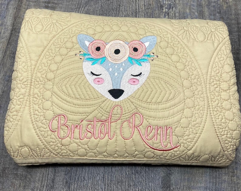K&ouml;nnte beinhalten: Eine beige gesteppte Babydecke mit einem wei&szlig;en Hirsch, der darauf gestickt ist. Der Hirsch tr&auml;gt eine rosa Blumenkrone und der Name "Bristol Renn" ist darunter gestickt.