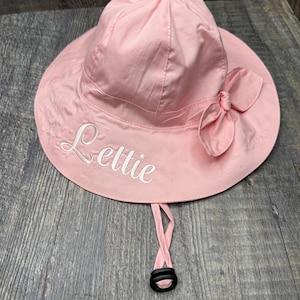 May include: A light pink sun hat with a wide brim and a decorative bow. The hat has the name "Lettie" embroidered in white script. It features a chin strap with a black adjustable toggle.