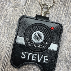 May include: Black leather keychain with a white and gray embroidered design of a speaker and the name "STEVE".