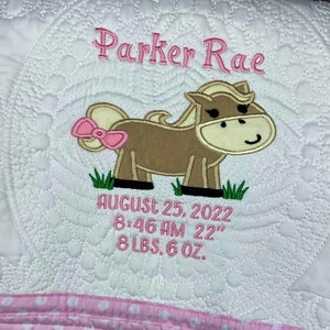 May include: A white quilted baby blanket with a pink and brown embroidered horse design. The blanket also includes the name "Parker Rae", the date "August 25, 2022", the time "8:46 AM", the length "22"
