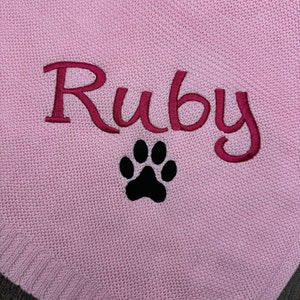 K&ouml;nnte beinhalten: Ein hellrosa Strickstoff mit dem eingestickten Namen Ruby in einer dunkelmagentafarbenen Schreibschrift, platziert direkt &uuml;ber einem einfarbig schwarzen, gestickten Pfotenabdruck. Die Textur des Strickmaterials ist auf der gesamten Fl&auml;che sichtbar.