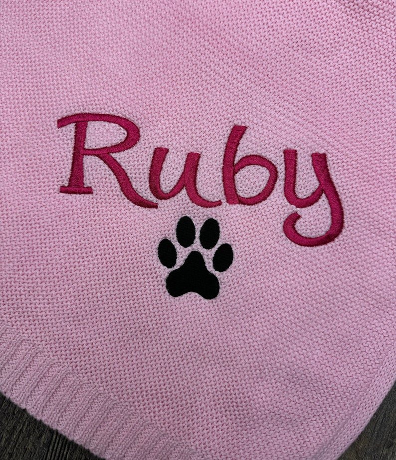 K&ouml;nnte beinhalten: Ein hellrosa Strickstoff mit dem eingestickten Namen Ruby in einer dunkelmagentafarbenen Schreibschrift, platziert direkt &uuml;ber einem einfarbig schwarzen, gestickten Pfotenabdruck. Die Textur des Strickmaterials ist auf der gesamten Fl&auml;che sichtbar.
