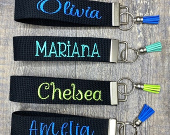 May include: Four black keychains with silver key rings and colorful tassels. Each keychain has a different name embroidered on it: Olivia, Mariana, Chelsea, and Amelia.