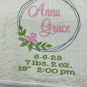 May include: Cream-coloured, quilted baby blanket with embroidered details. Features the name "Anna Grace" in pink script within a grey circle, accented with pink flowers and green leaves. Includes baby's birth date, weight (3.2 kg), and length (48 cm).