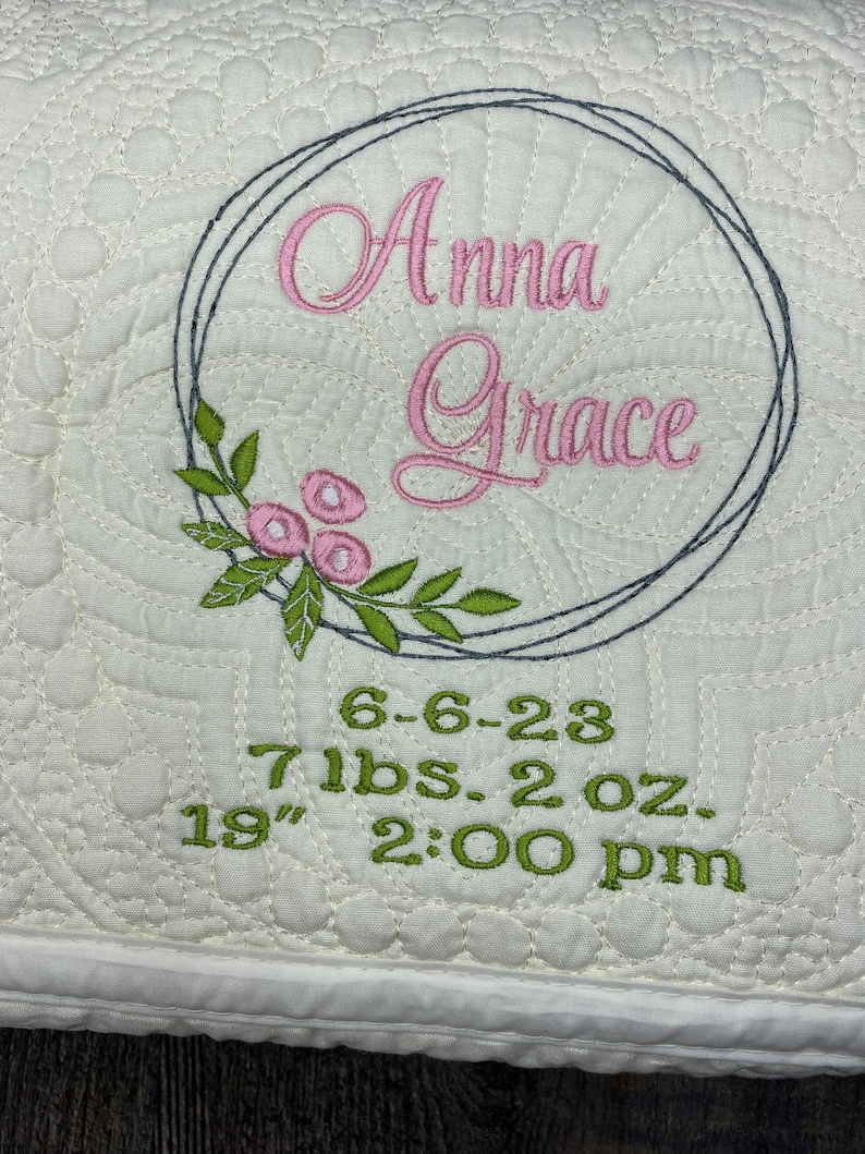 May include: Cream-coloured, quilted baby blanket with embroidered details. Features the name "Anna Grace" in pink script within a grey circle, accented with pink flowers and green leaves. Includes baby's birth date, weight (3.2 kg), and length (48 cm).