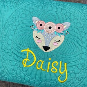 K&ouml;nnte beinhalten: T&uuml;rkise gesteppte Decke mit einem gestickten wei&szlig;en und rosa Reh mit einer Blumenkrone und dem Namen "Daisy" in Gelb gestickt.