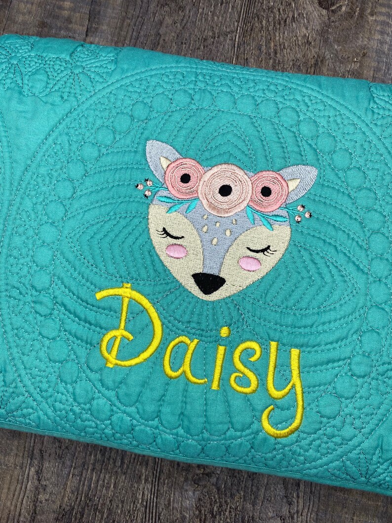 K&ouml;nnte beinhalten: T&uuml;rkise gesteppte Decke mit einem gestickten wei&szlig;en und rosa Reh mit einer Blumenkrone und dem Namen "Daisy" in Gelb gestickt.