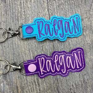 May include: Two personalized keychains with the name "Raegan" embroidered in purple thread. The top keychain has a light blue leather base and a purple metal ring. The bottom keychain has a purple leather base and a pink metal ring.
