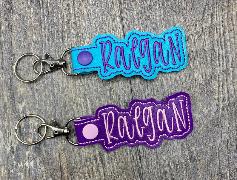 May include: Two personalized keychains with the name "Raegan" embroidered in purple thread. The top keychain has a light blue leather base and a purple metal ring. The bottom keychain has a purple leather base and a pink metal ring.