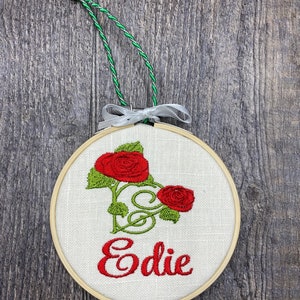 May include: A round, wooden-framed embroidery hoop with white linen fabric. The fabric features an embroidered design of two red roses with green leaves and stems. The name "Edie" is embroidered in red below the roses.