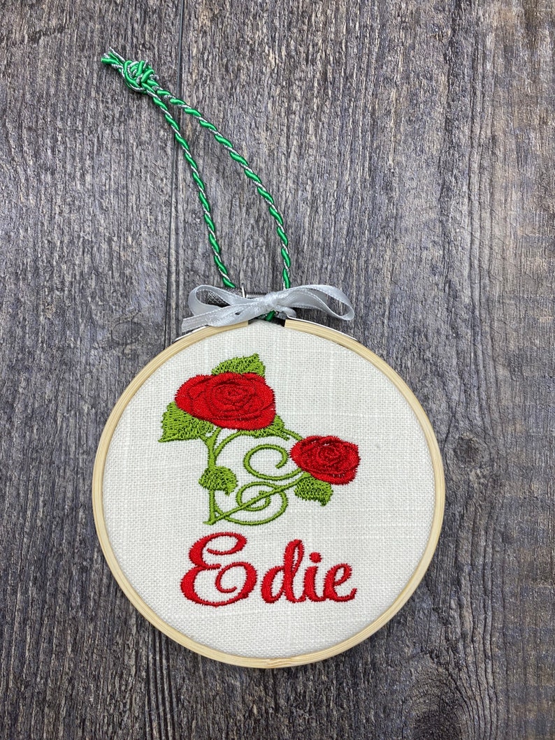 May include: A round, wooden-framed embroidery hoop with white linen fabric. The fabric features an embroidered design of two red roses with green leaves and stems. The name "Edie" is embroidered in red below the roses.