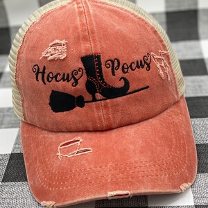May include: A distressed coral colored baseball cap with a black mesh back. The front of the cap features a black embroidered design of a witch's boot, broom, and the text "Hocus Pocus".