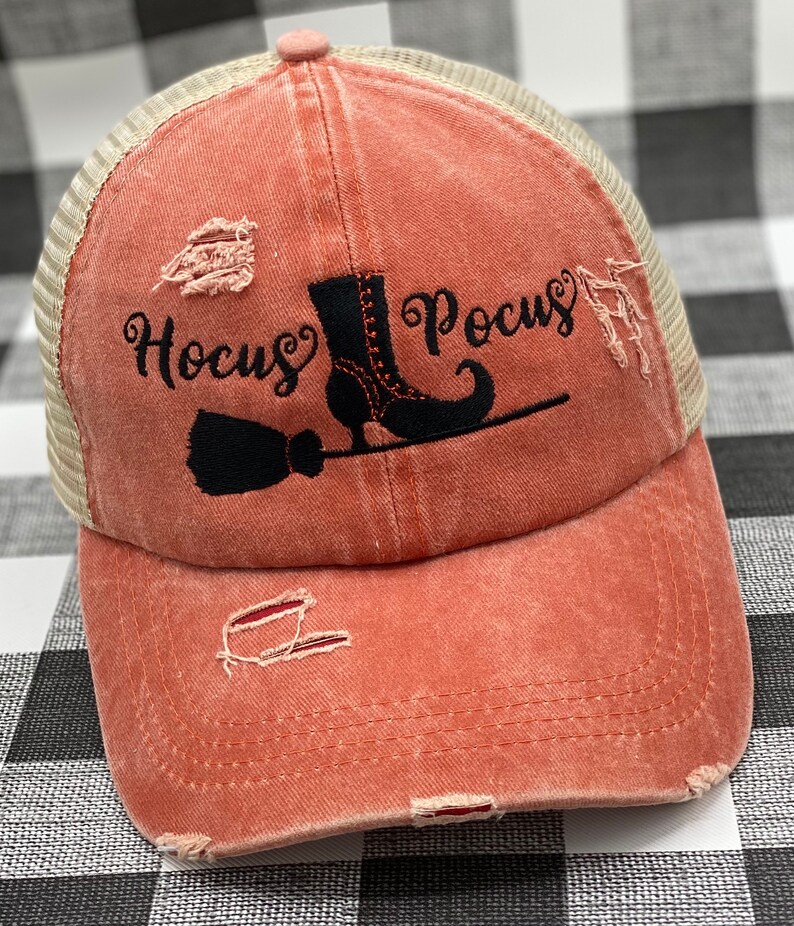 May include: A distressed coral colored baseball cap with a black mesh back. The front of the cap features a black embroidered design of a witch's boot, broom, and the text "Hocus Pocus".