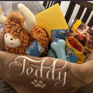 K&ouml;nnte beinhalten: Ein beigefarbener Strickkorb mit dem Namen Teddy und einer Pfotenabdruck-Stickerei in Wei&szlig;. Darin befinden sich ein orangefarbenes Hochlandrind-Pl&uuml;schtier, ein hellblaues knochenf&ouml;rmiges Spielzeug, ein blauer strukturierter Ball, ein gelber Umschlag und Leckerlis.