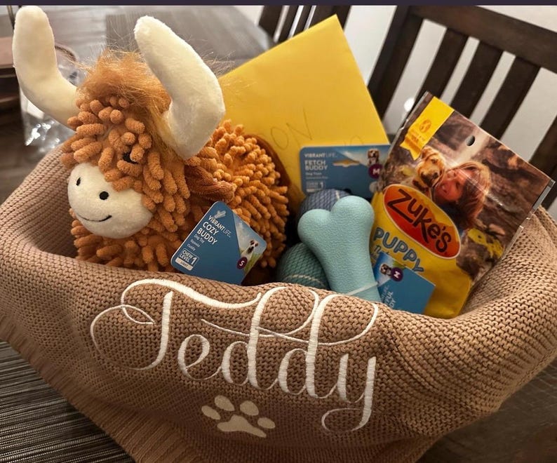 K&ouml;nnte beinhalten: Ein beigefarbener Strickkorb mit dem Namen Teddy und einer Pfotenabdruck-Stickerei in Wei&szlig;. Darin befinden sich ein orangefarbenes Hochlandrind-Pl&uuml;schtier, ein hellblaues knochenf&ouml;rmiges Spielzeug, ein blauer strukturierter Ball, ein gelber Umschlag und Leckerlis.