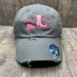 May include: A dark gray distressed baseball cap with a pink embroidered witch boot, broom, and the text "Hocus Pocus".