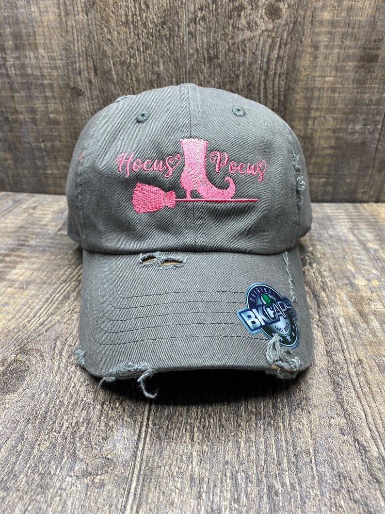 May include: A dark gray distressed baseball cap with a pink embroidered witch boot, broom, and the text "Hocus Pocus".