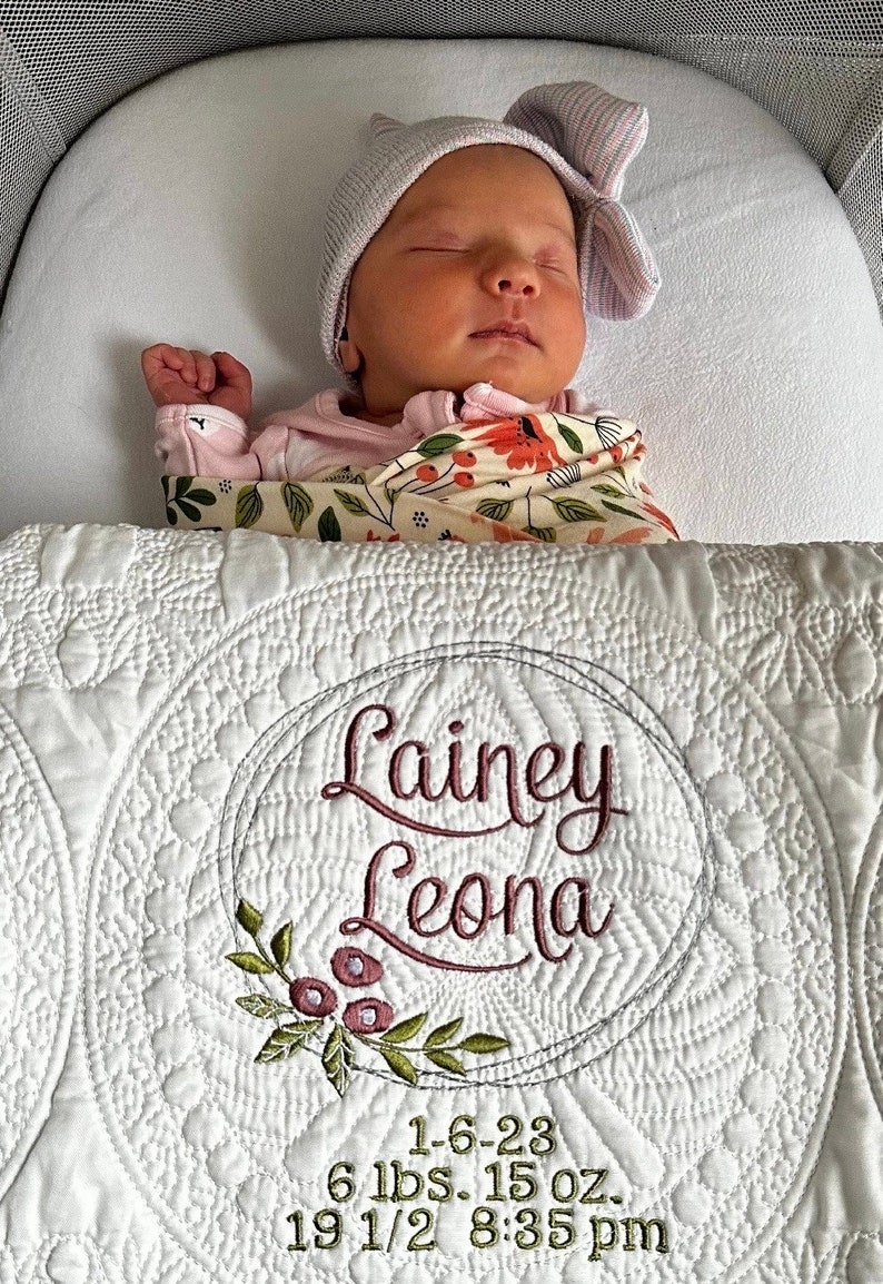May include: A sleeping newborn baby wearing a pink hat and floral blanket, in a bassinet. A white quilt with the name "Lainey Leona" embroidered in cursive, and birth details, is visible.