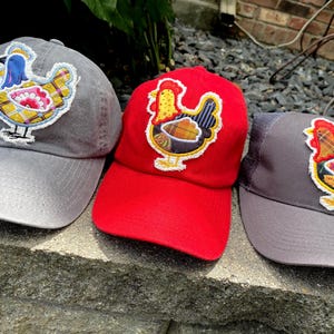 May include: Three baseball caps with different colored fabric and embroidered chicken designs. The caps are gray, red, and gray with a mesh back.