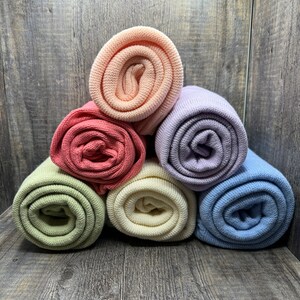 K&ouml;nnte beinhalten: Eine Sammlung von sechs gerollten Strickstoffen in Pastell- und gedeckten T&ouml;nen, darunter Pfirsich, Lavendel, Hellblau, Salbeigr&uuml;n, Creme und Altrosa. Jedes St&uuml;ck weist eine gleichm&auml;&szlig;ige, fein gestrickte Textur auf, die in einer Pyramidenform gestapelt ist.