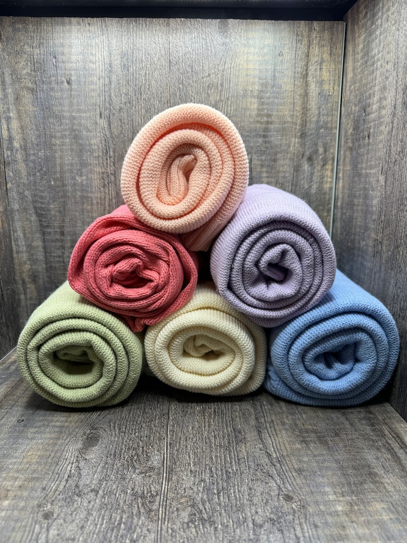 K&ouml;nnte beinhalten: Eine Sammlung von sechs gerollten Strickstoffen in Pastell- und gedeckten T&ouml;nen, darunter Pfirsich, Lavendel, Hellblau, Salbeigr&uuml;n, Creme und Altrosa. Jedes St&uuml;ck weist eine gleichm&auml;&szlig;ige, fein gestrickte Textur auf, die in einer Pyramidenform gestapelt ist.