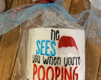 May include: A white toilet paper roll with red and blue embroidery that reads "He sees you when you're pooping". The roll is wrapped in clear plastic with a blue and red ribbon.