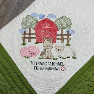 May include: A white quilted baby blanket with a green border. The center of the blanket features an embroidered scene of a red barn, a horse, a pig, and a sheep. The text "To Isaac Michael From Mamaw" is embroidered below the scene.