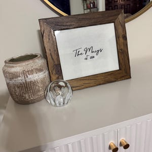 May include: A rectangular wooden picture frame containing a white insert with the black text The Macys est. 2024, positioned next to a textured beige ceramic vase and a clear glass paperweight with internal white swirling patterns.