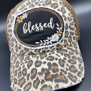 May include: A brown and white leopard print trucker hat with a black oval patch that says "blessed" in white embroidery. The patch is surrounded by white embroidered flowers.