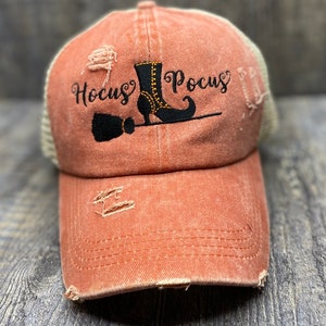 May include: A distressed orange and beige baseball cap with an embroidered witch boot, broom, and the text "Hocus Pocus" in black thread.