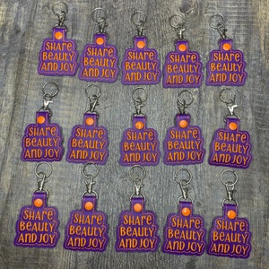 May include: Fifteen purple keychains with silver key rings. Each keychain has a white stitched border and features the text "Share Beauty and Joy" in orange lettering.