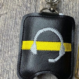 May include: A black faux leather keychain holder featuring a horizontal yellow embroidered stripe and a silver embroidered headset icon. The rectangular holder has rounded corners, black stitching along the edges, and a silver metal lobster clasp.