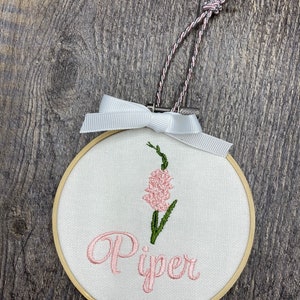 May include: A white embroidery hoop with a pink flower and the name "Piper" embroidered in pink.