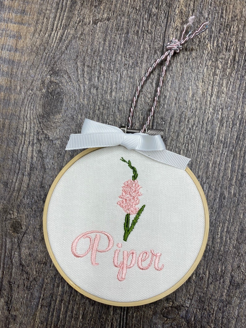 May include: A white embroidery hoop with a pink flower and the name "Piper" embroidered in pink.