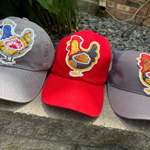 May include: Three baseball caps with embroidered chicken designs. The caps are in gray and red. The chicken designs feature a variety of colors, including yellow, blue, red, and brown. The caps are sitting on a stone surface.