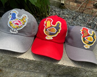 May include: Three baseball caps with embroidered chicken designs. The caps are in gray and red. The chicken designs feature a variety of colors, including yellow, blue, red, and brown. The caps are sitting on a stone surface.