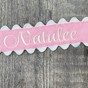 May include: A pink ribbon pacifier clip with the name "Natalee" embroidered in white script. The ribbon is edged with white scalloped trim and has a silver-colored metal clip and loop.