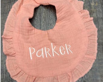 May include: A peach-colored baby bib with a ruffled edge and a white button closure. The bib is personalized with the name "Parker" embroidered in white.