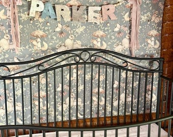 May include: A baby crib with a black metal frame and a floral patterned crib sheet. A banner spelling "PARKER" hangs above the crib. The banner is made of various patterned fabrics and colors, with pink ribbons on each side.