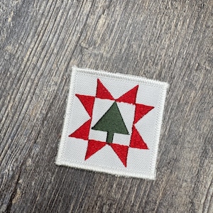 May include: A square embroidered patch featuring a white background with a red eight-pointed star design. Centered within the star is a solid dark green triangle representing a tree. The patch has a clean, stitched border and a flat texture.