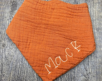 May include: An orange, triangular baby bib with the name "Mack" embroidered in white thread. The bib is made of a textured fabric and is laid flat on a wooden surface.