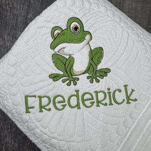 May include: White quilted blanket with a green frog embroidery and the name "Frederick" in green. The blanket features a detailed quilted pattern and a whimsical frog design, perfect for a nursery or child's room.