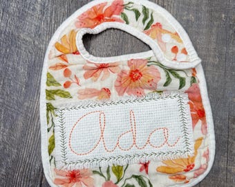 May include: Floral baby bib with the name "Ada" embroidered in orange thread. The bib features a white border and a floral pattern with orange, pink, and green flowers. The bib is made of soft fabric and has a snap closure.