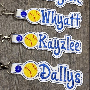 May include: Six white keychains with blue embroidered names and a yellow softball graphic. The names are Izzy, Layton, Wyatt, Kayzlee, Dallys, and Omaria.