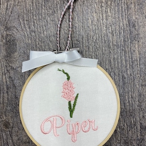 May include: A white embroidery hoop with a pink flower and the name "Piper" embroidered in pink thread.