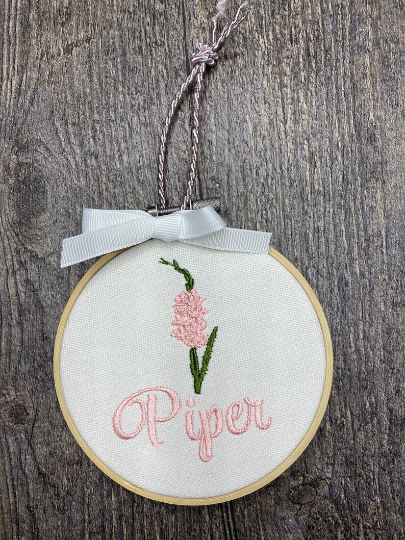 May include: A white embroidery hoop with a pink flower and the name "Piper" embroidered in pink thread.