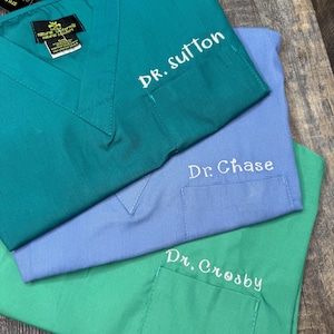 May include: Three teal, periwinkle, and green medical scrubs with embroidered names. The teal scrub is labeled "Dr. Sutton", the periwinkle scrub is labeled "Dr. Chase", and the green scrub is labeled "Dr. Crosby". Each scrub has a chest pocket.