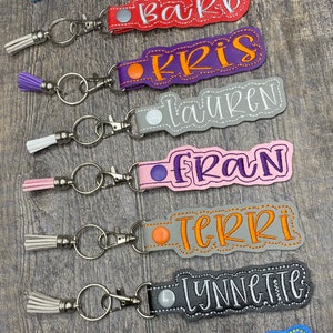 May include: Seven colorful keychains with different names embroidered on them. Each keychain has a small tassel and a silver key ring. The names are: Kit, Barb, Kris, Lauren, Fran, Terri, Lynnette, and Trace.